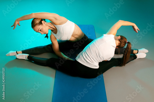 Beautiful two young slim gymnast women in sports clothing stretching on the floor on fitness mat in neon lights. Flexible muscular women doing gymnastic exercise.