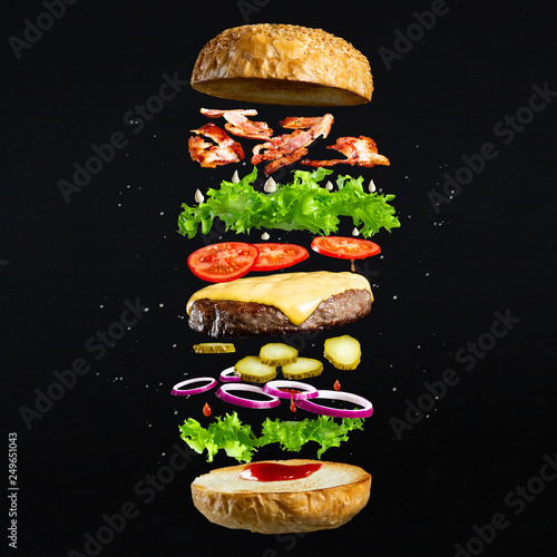 Floating burger isolated on black wooden background. Ingredients of a delicious burger with ground beef patty, lettuce, bacon, onions, tomatoes and cucumbers