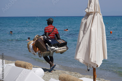 Vendor with counterfeit products on the beach in Chaniotis, Greece. Street vendor selling popular brands replicas bags by a Greek beach shore on a sunny summer day.