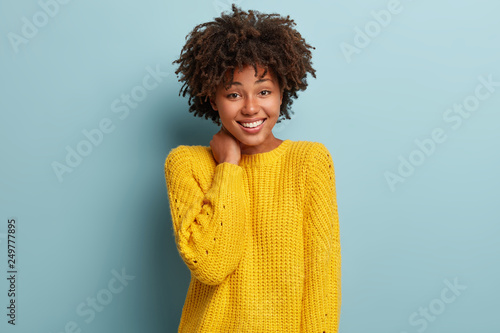 Happy impressed young woman with dark skin, toothy smile, dressed in casual yellow jumper, keeps hand on neck, expresses good feelings, recieves compliment from handsome man. Emotions concept