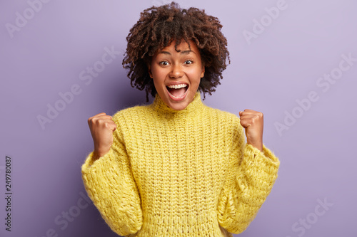 Overjoyed carefree African American woman raises clenched fists, shows success gesture, triumphs over purple background, wears knitted winter yellow sweater, isolated over purple background.