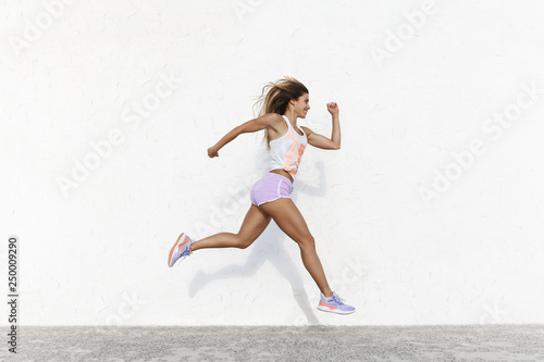 Strong athletic happy sportswoman wearing sport shorts jump across white concrete wall facing forward running in air, smiling accomplished, setting goal, workout, jogging, sprint along quay