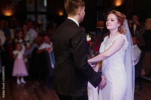 Romantic bride and groom dancing and holding hands at wedding reception in restaurante, newlywed couple first dance at evening party, guests looking in the background