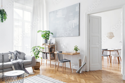 Stylish scandinavian open space with design furniture, plants, bamboo bookstand and wooden desk. Brown wooden parquet. Abstract painting. Modern decor of bright room next to dining room. 