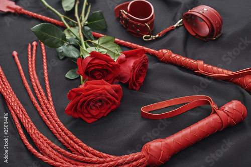 Playing BDSM games. Top view of bdsm leather kit (handcuffs, whip) and roses against of black silk fabric