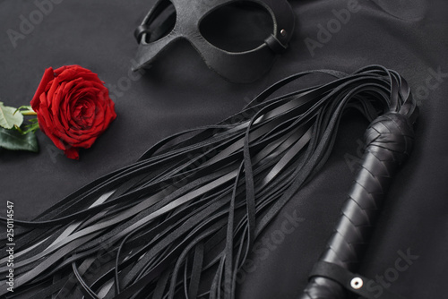 Role playing. Top view of bdsm leather kit ( black whip and mask) with red rose on black silk