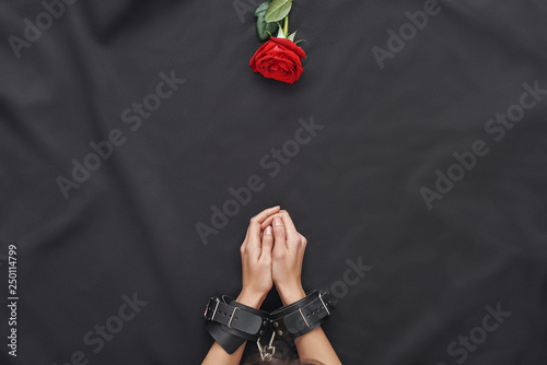 Explore your sexual fantasies. Top view of female hands wearing black leather handcuffs and red rose against of black silk