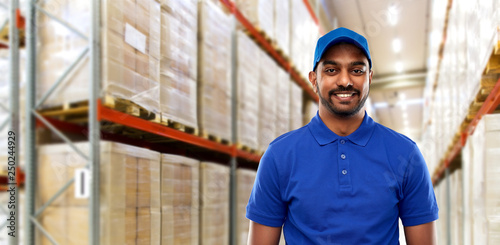logistic business, shipment and people concept - happy indian delivery man or warehouse worker in blue uniform over goods background