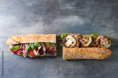 Two Beef baguette sandwiches with champignon mushrooms, green salad, fried onion, tomatoes over blue texture background. Flat lay, space