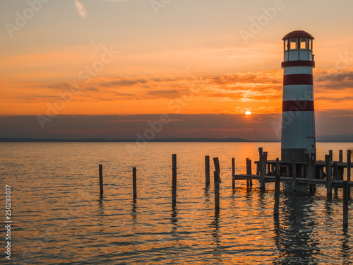 "White and red striped Lake's lantern in Neusiedlersee, on Austrian-Hungarian border. Sun sets over the horizon in the clouds. Sky is orange, lake surface is soft and tender. Few clouds on the sky. "