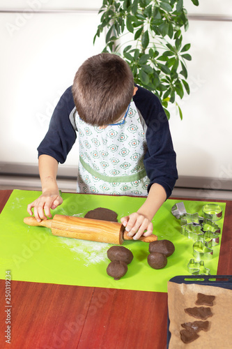Chłopiec ubrany w fartuch kuchenny wałkuje brązowe ciasto drewnianym wałkiem na silikonowej zielonej stolnicy. Dziecko w kuchni.
