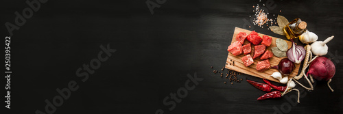 Uncooked fresh diced beef meat with herbs and oil on an old rustic wooden kitchen board over black background. Top view with free space for text. Banner