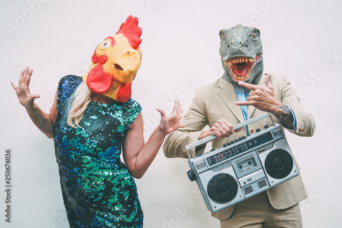 Crazy senior couple wearing chicken and t-rex mask while dancing outdoor - Mature trendy people having fun celebrating and listening music with boombox - Absurd concept of masquerade funny holidays