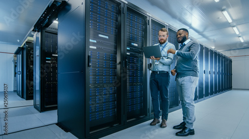 Bearded IT Technician in Glasses with a Laptop Computer and Black Male Engineer Colleague are Talking in Data Center while Working Next to Server Racks. Running Diagnostics or Doing Maintenance Work. 