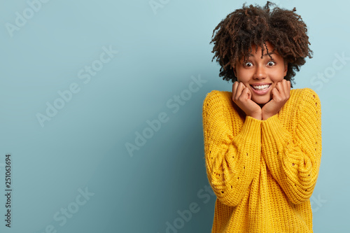 Emotional thrilled positive dark skinned lady shows teeth, smiles and has surprised joyful expression, listens something with great interest, dressed in yellow casual sweater, isolated on blue wall