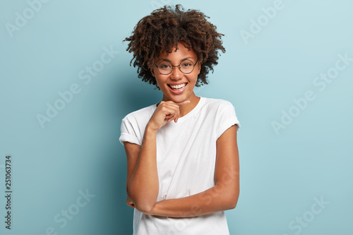 Portrait of cheerful black woman with Afro hairstyle, smiles gently, imagines something pleasant, wears round glasses and t shirt models against blue background. Emotions and human expressions concept