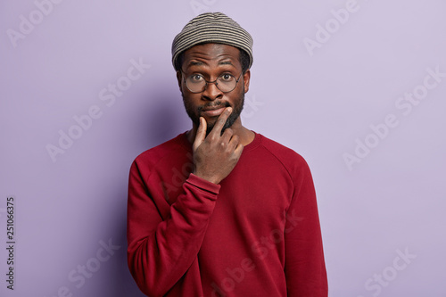 Stylish ethnic unshaven man holds chin, has surprised expression, wonders what to do in difficult situation, wears hat, spectacles and red jumper, isolated over purple background, has hesitant look