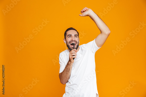 Photo of optimistic man 30s listening to music using earphones and mobile phone, isolated over yellow background
