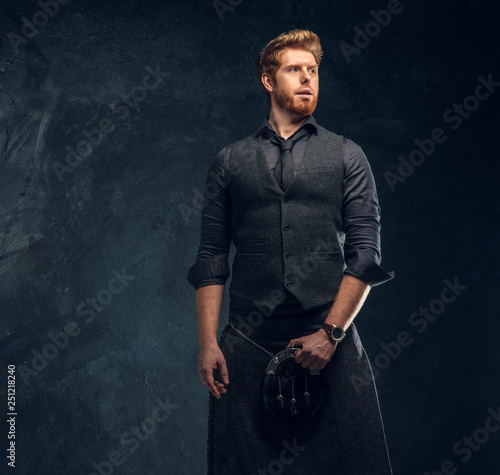 Portrait of a handsome redhead man dressed in an elegant vest with tie and kilt in studio against a dark textured wall