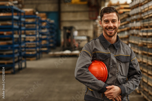 Shot of a handsome young bearded factory worker in uniform holding protective hardhat smiling joyfully to the camera posing at the warehouse of a metalworking company