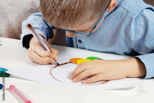 Chłopiec w niebieskiej koszuli i okularach siedzi przy biurku i w skupieniu wykonuje pracę plastyczną z kolorowego papieru. Chłopiec maluje brązowym flamastrem.