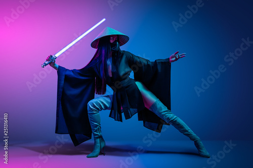 Concept on cosmic cosplay. Сontemporary portrait a young athletic woman in traditional Japanese black kimono, an Asian hat and highboots is holding a lightsaber and posing on neon blue-pink background