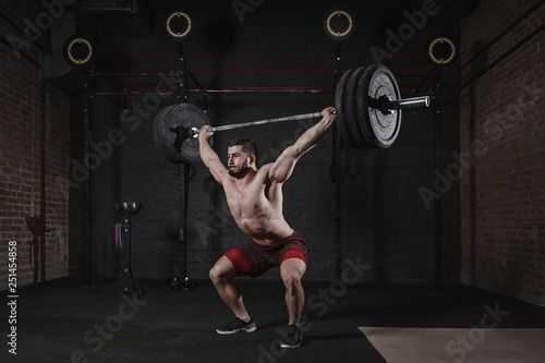 Young crossfit athlete lifting heavy barbell overhead at the gym. Handsome man doing functional training. Practicing powerlifting.