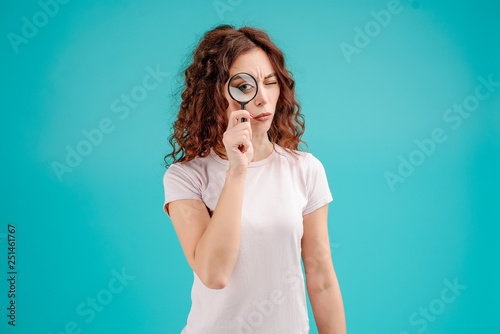 Attractive brunette girl with curly hair isolated over blue turquoise background looking at something through magnifying glass with a big eye. Vision concept