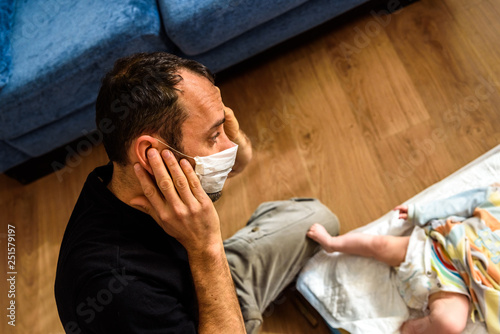 Novice father changing the smelly diaper of a baby, wearing a mask for bad smells with a gesture of disgust.