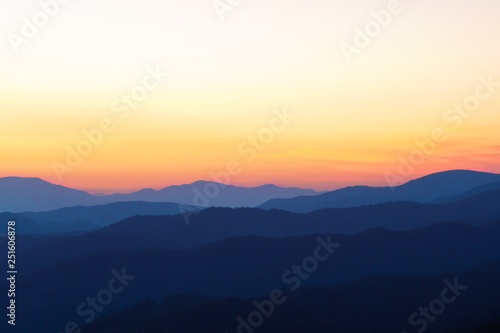 Panorama of the peaks of beautiful mountains covered with trees at sunset on a sunny day in summer. Orange glow on the sky after full sunset. Dark silhouettes of mountains. Solar reflection