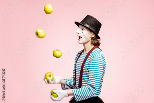 Pantomime with white facial makeup juggling with apples on the pink background in the studio