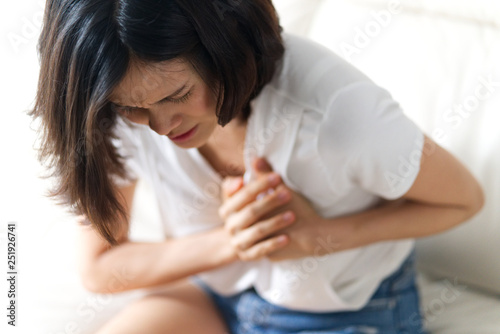 Unwell condition and sickness concept; Asian woman having heart attack sitting on sofa. She puts both hand on her left chest feeling painful and need emergency medical assistance.