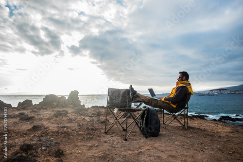 Digital nomad and traveler middle age adult man sitting and working with internet and laptop in totally fredom in front of the ocean - backpacker lifestyle and alternative office for happy life