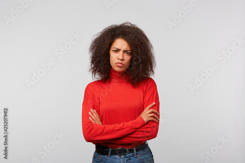 Serious suspicious latino woman with curly afro hair standing with crossed hands and frowning, expressing distrustfulness and disappointment, over white background