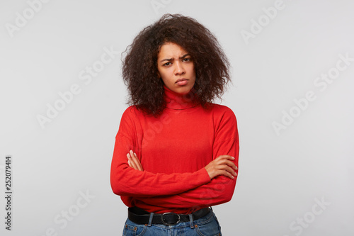 Indoor photo of serious suspicious latino woman with curly afro hair standing with crossed hands and frowning, expressing distrustfulness and disappointment, over white background