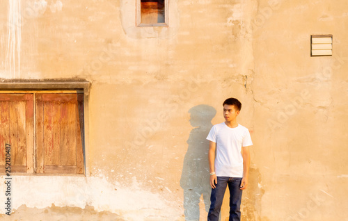 A young Thai boy is standing front of the dirty wall in the white shirt with moody face