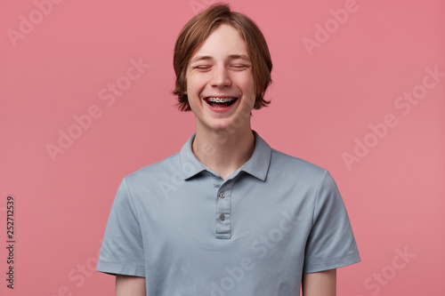 Close up of nice blue-eyed neatly combed young guy with braces on teeth joyfully laughing closed his eyes of fun wears polo t-shirt looks happy isolated over pink background
