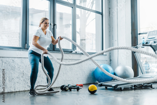 cheerful overweight woman training with battle ropes in gym