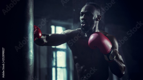 Hands of boxer over dark gym background. Strength, attack and motion concept. Fit african american model in movement. Naked muscular athlete in red gloves. Sporty man during boxing