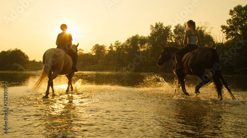 LENS FLARE: Active girls riding their strong horses down the river shallows.