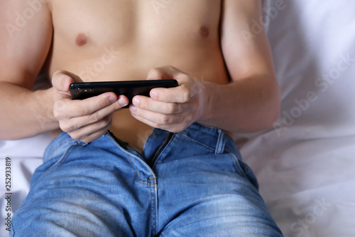 Smartphone in male hands, man in unzipped jeans with naked torso relaxing in bed. Concept of watching video, playing mobile games at home, sex dating, online communication, video chat