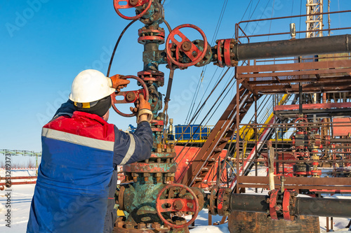 An employee of an oil company performs work on the well. Rotates the steering wheel on the fountain fittings. In the background equipment for oil production. Winter day at the polar oil field.