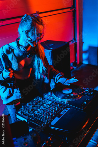 happy blonde dj woman holding bottle while touching vinyl record in nightclub