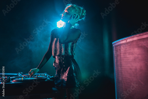 happy dj woman with blonde hair listening music and touching headphones in nightclub with smoke