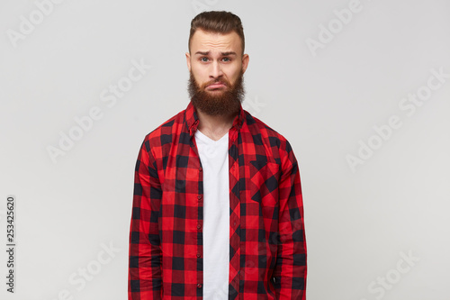 Emotional bearded man looks unhappy unsatisfied puzzled with incomprehension expresses question, wears shirt, stands isolated over white background.