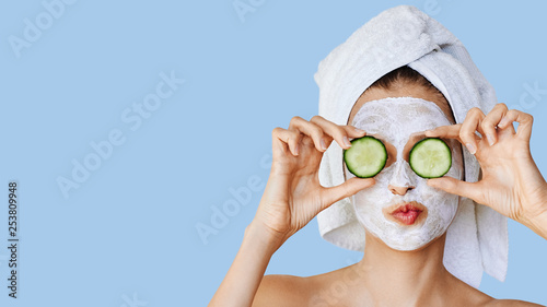Beautiful young woman with facial mask on her face holding slices of cucumber. Skin care and treatment, spa, natural beauty and cosmetology concept.