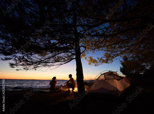 Romantic supper on lake shore at night. Tourist tent under trees and young family, man and woman at campfire on dark blue evening sky and clear water background. Tourism and camping concept.