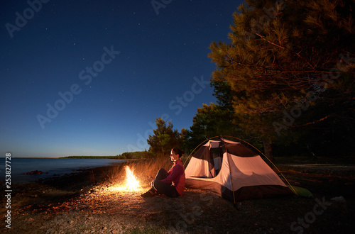 Night camping on lake shore. Happa attractive woman sitting relaxed in front of tent at campfire under bright starry sky on clear blue water and green forest background. Active lifestyle concept.