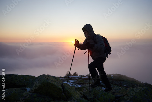Tourist hiker in warm jacket with backpack and trekking sticks walking on rocky mountain peak on copy space background of foggy misty landscape, clear blue sky and raising bright orange sun at dawn.