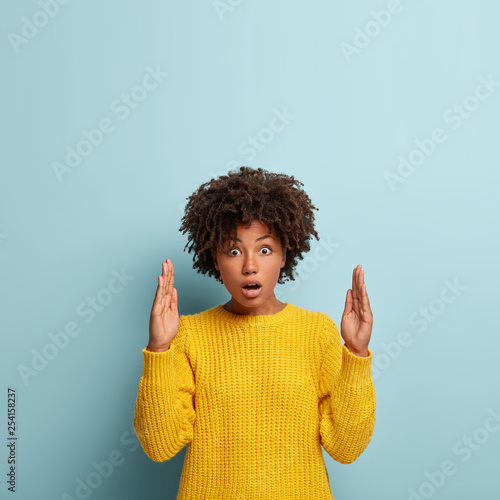 Pretty surprised impressed African American woman demonstrates something big, shapes huge objects with both hands, gestures actively, wears yellow sweater, isolated over blue wall, blank space